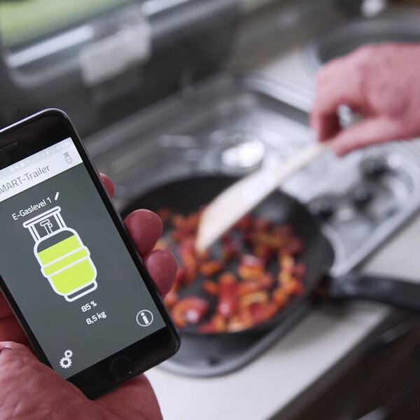 Eine Hand hält Smartphone auf dem in der App der Füllstand einer Gasflasche angezeigt wird, während die andere Hand im Hintergrund Essen zubereitet.