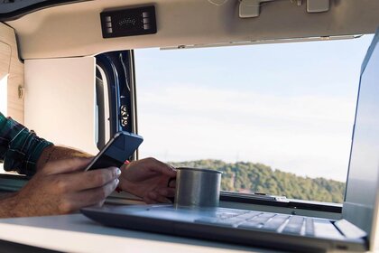 SMV Download. Ein Laptop auf einem Wohnmobiltisch. Rechte Hand hält ein Smartphone, die linke eine Blechtasse. Hintergrund Blick auf Baumlandschaft.
