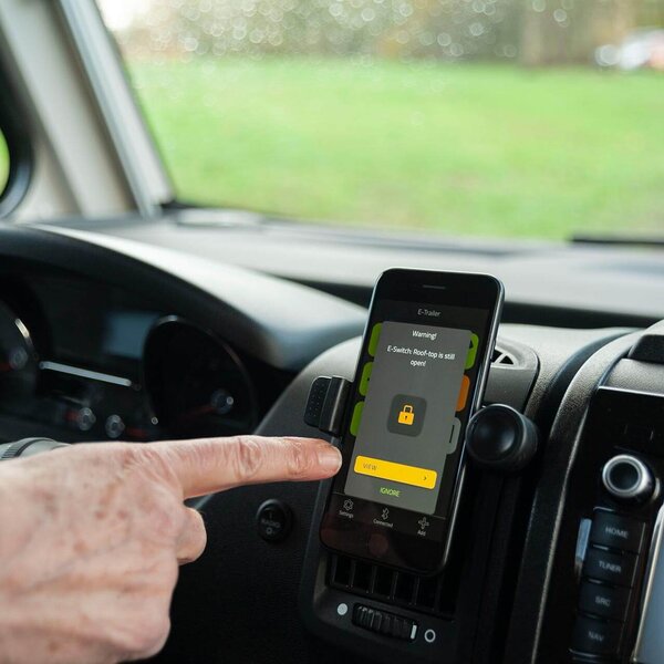 Hand zeigt im Cockpit eines Wohnmobils auf Smartphone mit E-Switch Warnmeldung.